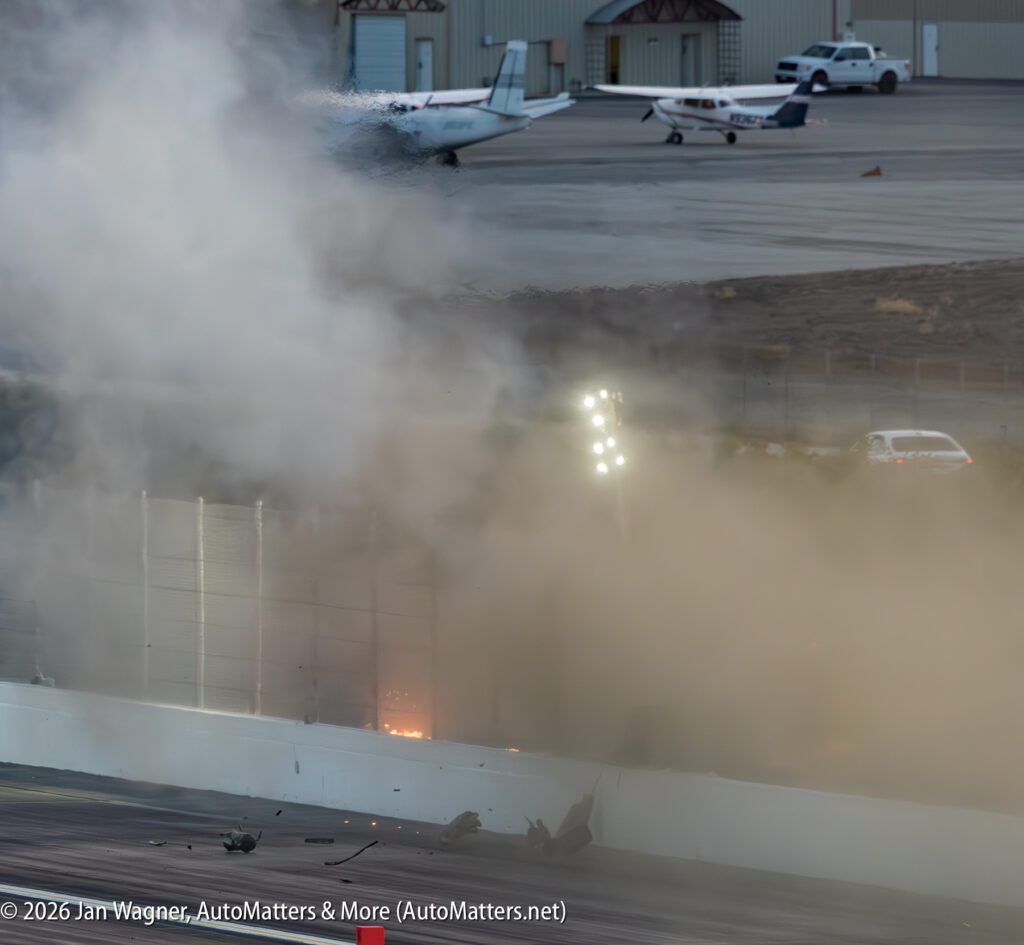 A large cloud of smoke and debris rises from a crash at a racetrack, with flames visible near the barrier and airplanes parked in the background.