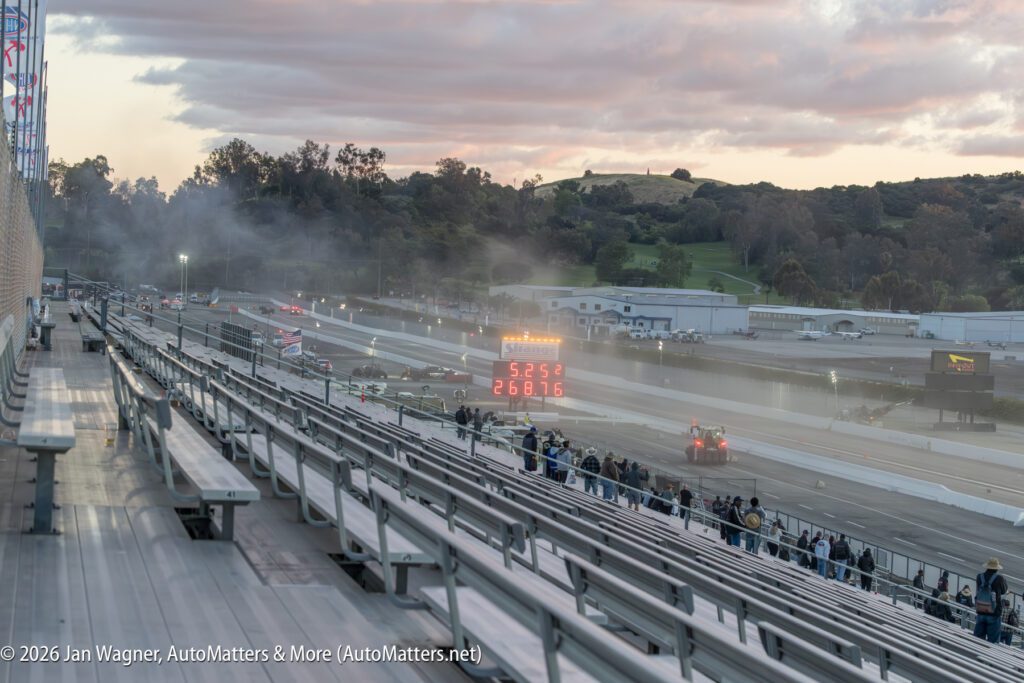 Drag racing cars speed down a track as digital display shows race times; spectators watch from grandstand under cloudy sky.