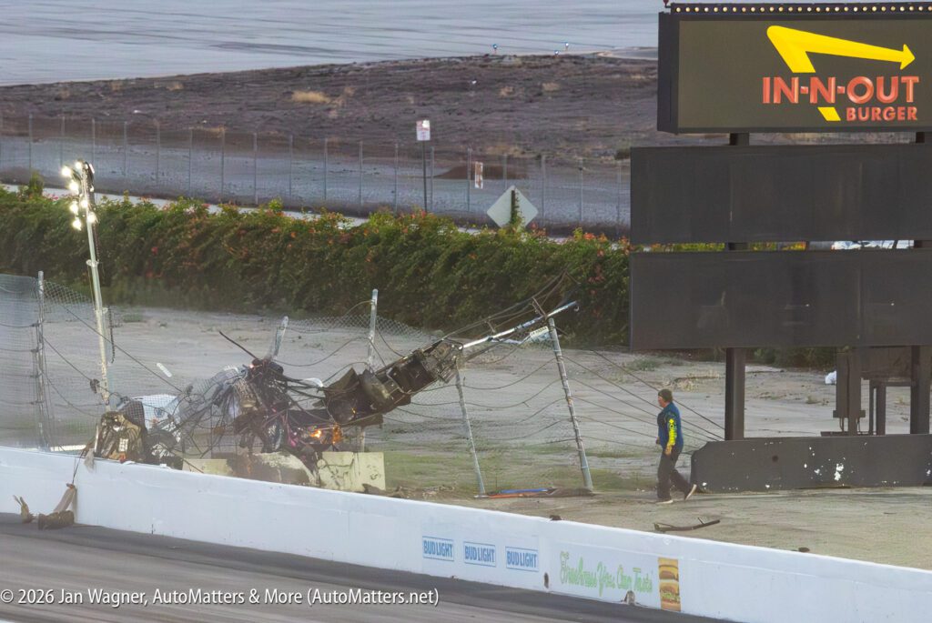 Damaged race car parts tangled in safety fencing beside a track barrier, with a person in safety gear walking nearby and an In-N-Out Burger sign in the background.