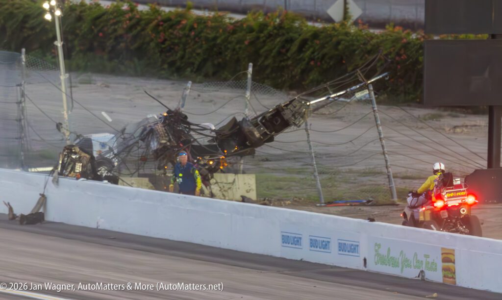 A damaged utility pole and debris lie on a racetrack while emergency responders examine the scene and a responder on a motorcycle looks on.