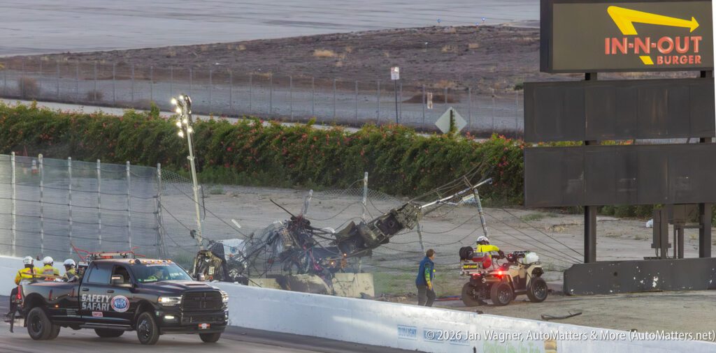 Safety personnel attend to a race track crash scene with a damaged safety fence, emergency vehicles, and debris scattered near an In-N-Out Burger sign.