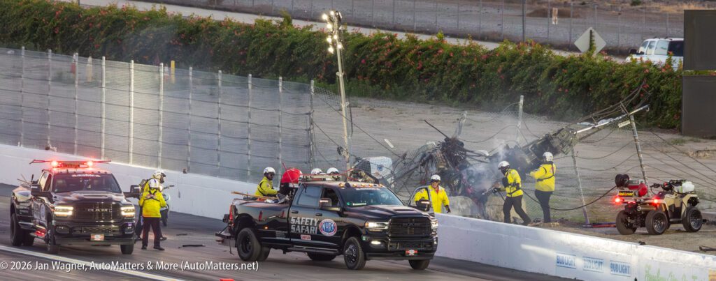 Emergency and safety crews respond at a racetrack where a crashed car has heavily damaged the safety fence, with trucks and personnel present at the scene.