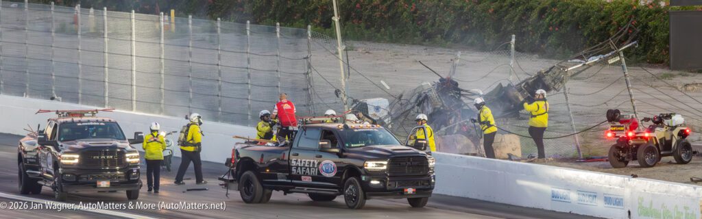 Safety crew and vehicles respond to a crash scene at a racetrack, with damaged barriers and debris visible in the background.