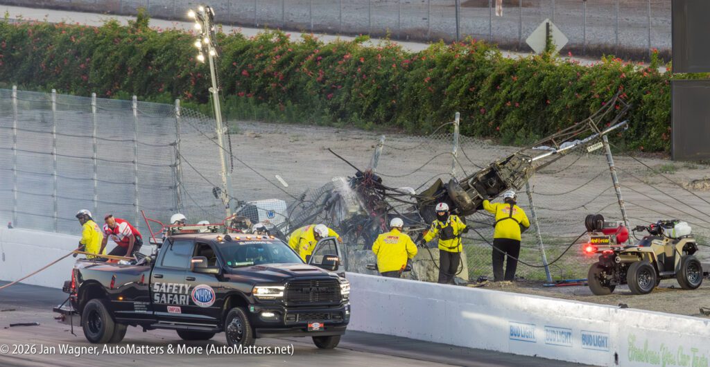 Safety crew attends to a race car that has crashed and become entangled in the fence at a racetrack, with emergency vehicles and personnel present.