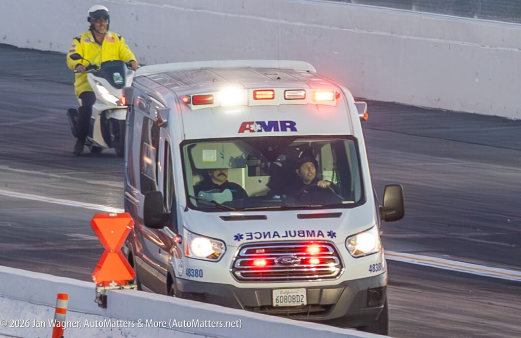 An ambulance with flashing lights drives on a racetrack, followed by a person in yellow gear riding a scooter.