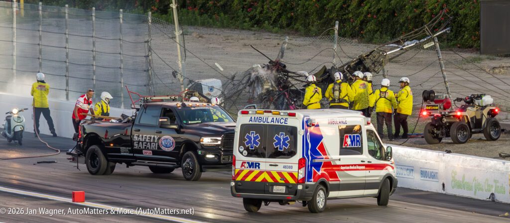 Crash scene at a racetrack with emergency personnel, an ambulance, a safety truck, and damaged fencing in the background.