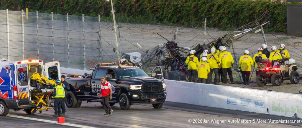 Emergency crews respond to a crash scene at a racetrack, attending to wreckage and preparing a stretcher near ambulances and a safety truck.