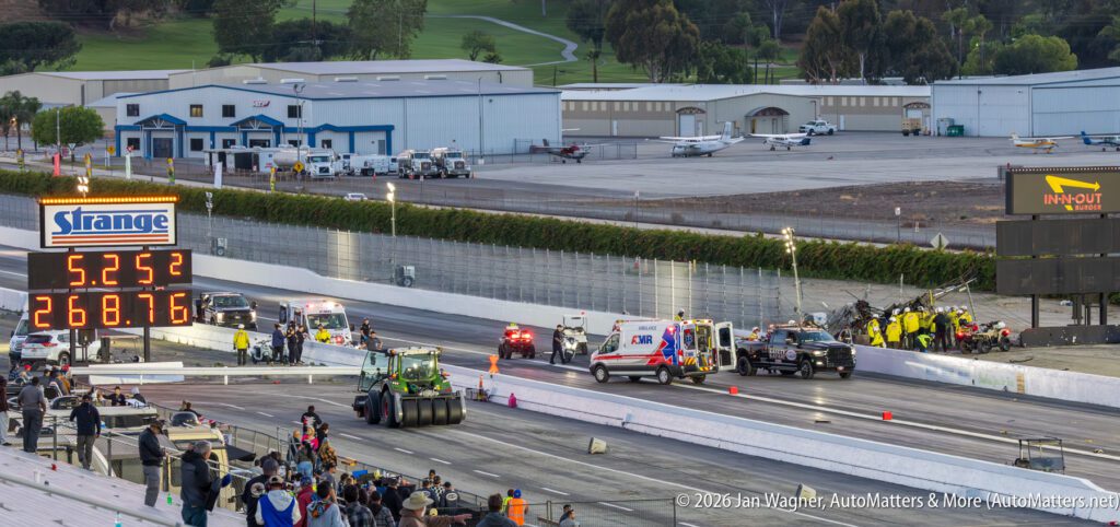 A drag racing track with emergency vehicles and crew attending to an incident; scoreboard displays elapsed time and speed; small airplanes visible in the background.