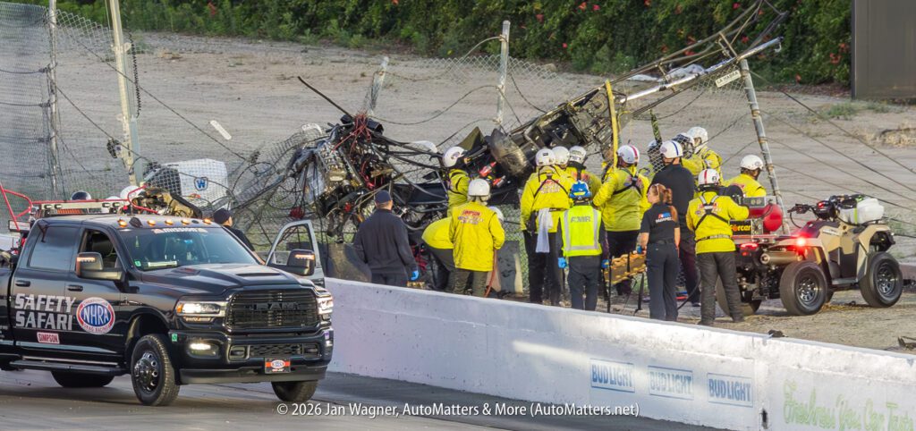 A group of safety crew members stands near a crashed race car entangled in a fence, with emergency vehicles parked nearby on a racetrack.