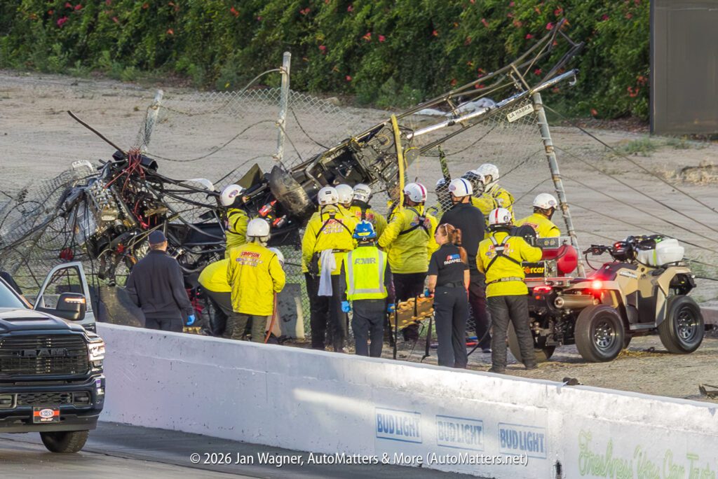 A group of emergency responders attends to a crashed race car that is heavily damaged and caught in a fence at a racetrack.