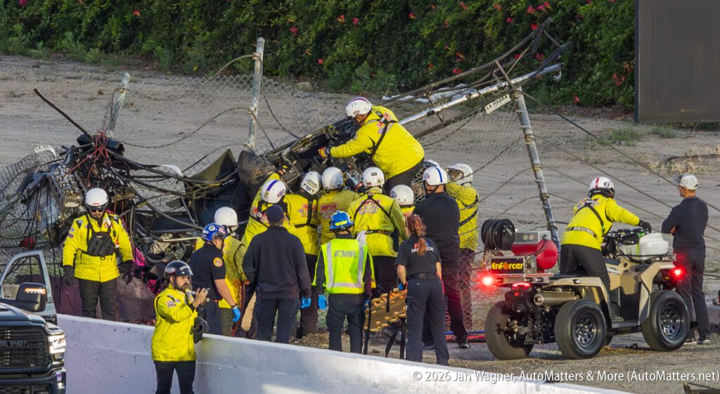 Emergency personnel attend to a crash scene with a damaged fence and vehicle debris on a racetrack, surrounded by various safety crew members and vehicles.