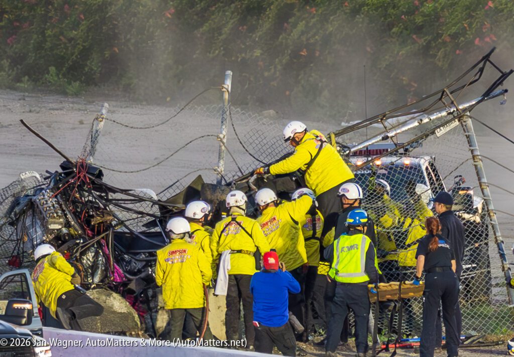 Emergency responders in yellow jackets work at the scene of a serious crash involving a damaged fence and vehicle debris, using tools to access the wreckage.
