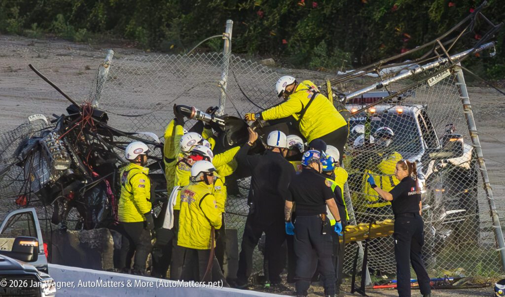 Emergency responders surround a damaged race car entangled in a fence at a racetrack, working to assist the driver following a crash.
