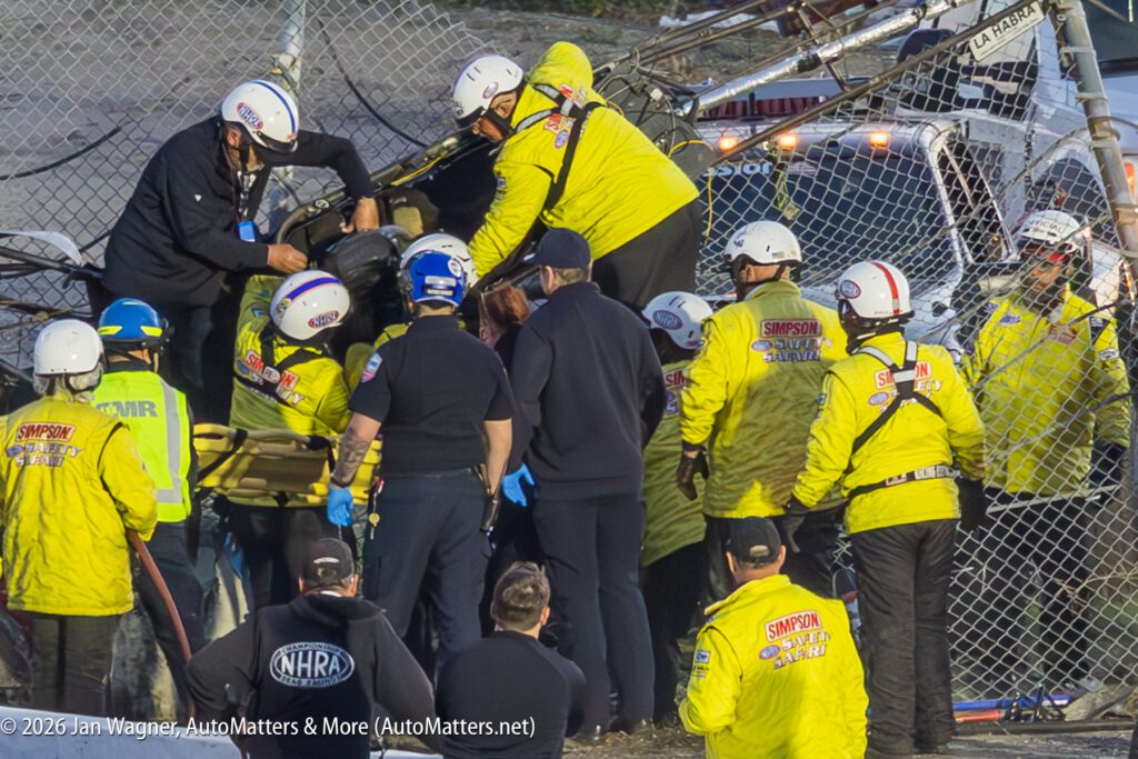 Emergency personnel in yellow and black uniforms assist at the scene of a crash involving a vehicle against a fence, with several responders attending.