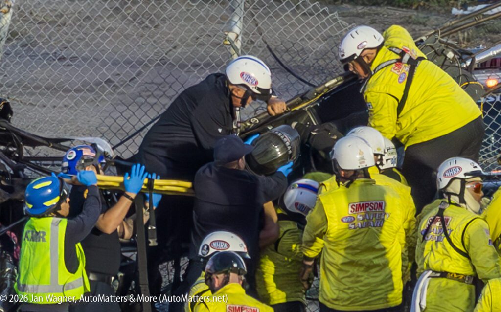 Rescue crew surrounds and assists a person in racing gear next to a damaged fence, with emergency personnel in yellow jackets and helmets.