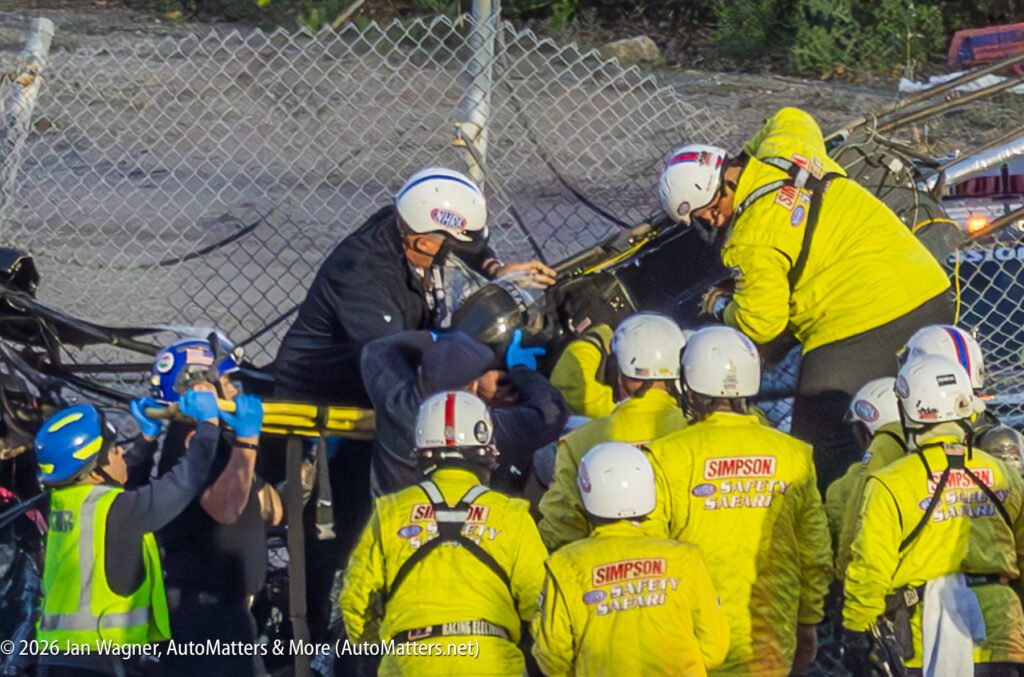 Safety crew in yellow suits assists and extracts a person from a damaged crash site near a chain-link fence, with bystanders and emergency staff observing.