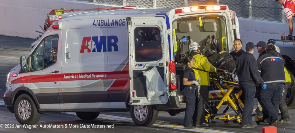 Paramedics load an individual on a stretcher into an AMR ambulance at a racetrack, surrounded by emergency personnel.