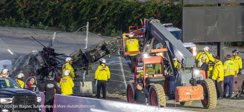 A crash scene at a racetrack shows emergency crews and a forklift removing wreckage from a fence, with safety personnel assessing the situation.