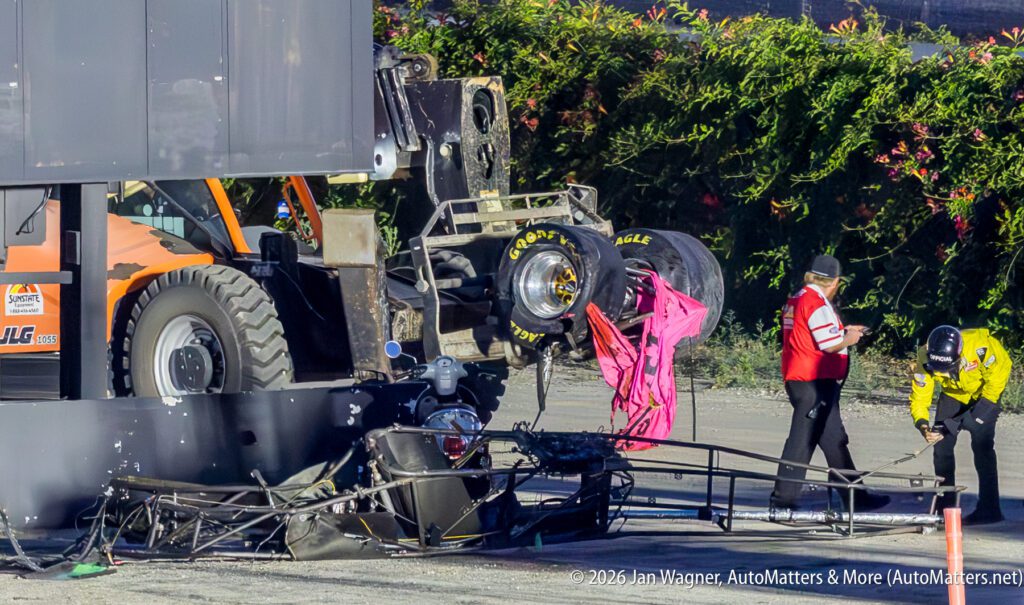 A crashed race car with detached parts lies near an orange construction vehicle as two officials assess the scene.