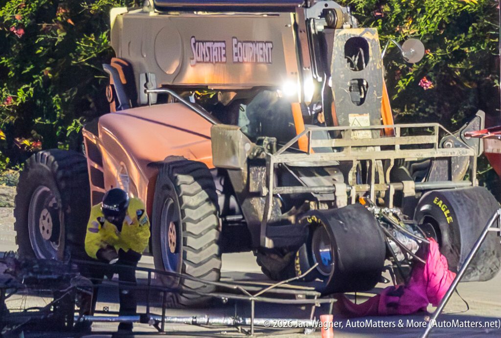 A safety worker and a wrecked vehicle are next to a large construction forklift on a race track, with car parts and equipment scattered on the ground.