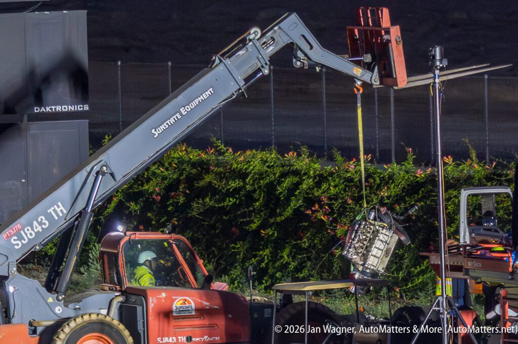A telescopic forklift lifts an engine block at night during an outdoor event, with workers and equipment visible around the work area.