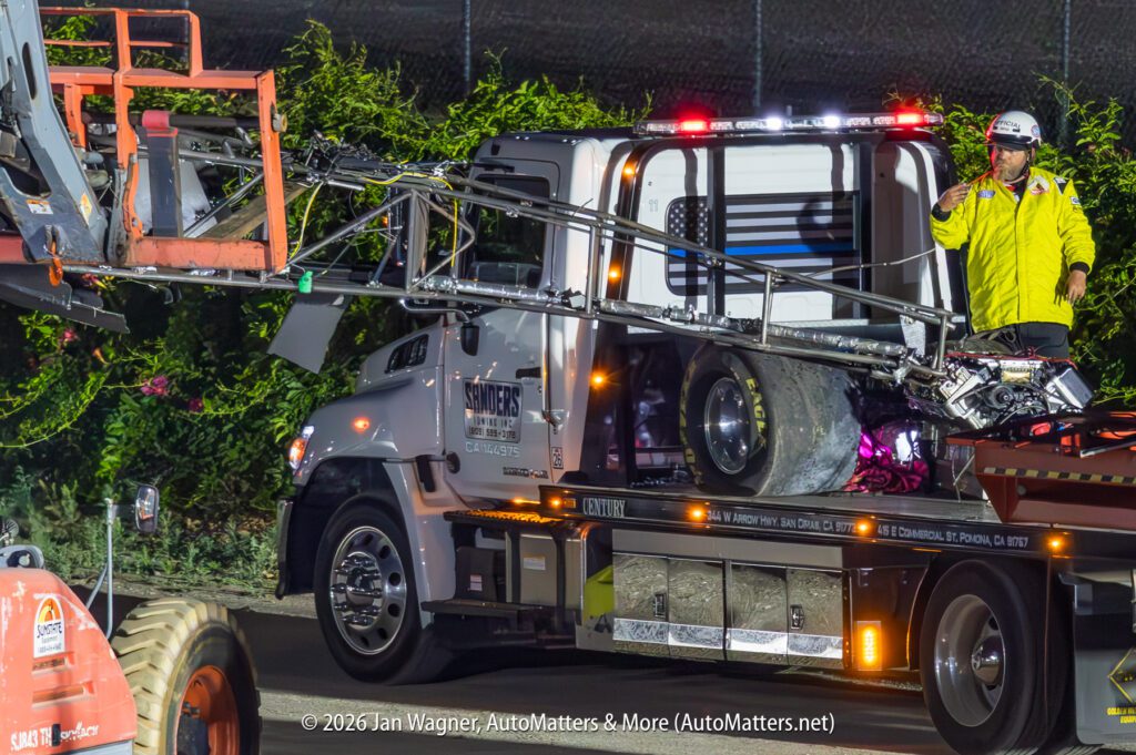 A tow truck transports a damaged motorcycle and a section of guardrail at night, while a worker in a yellow jacket stands nearby.