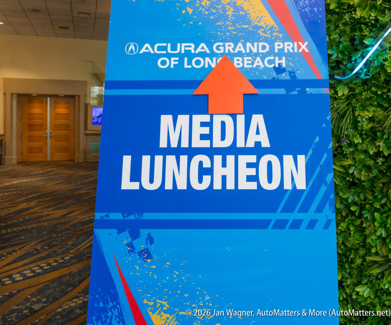 Sign for "Acura Grand Prix of Long Beach" Media Luncheon with an arrow pointing up, displayed indoors on a patterned carpet near double doors.