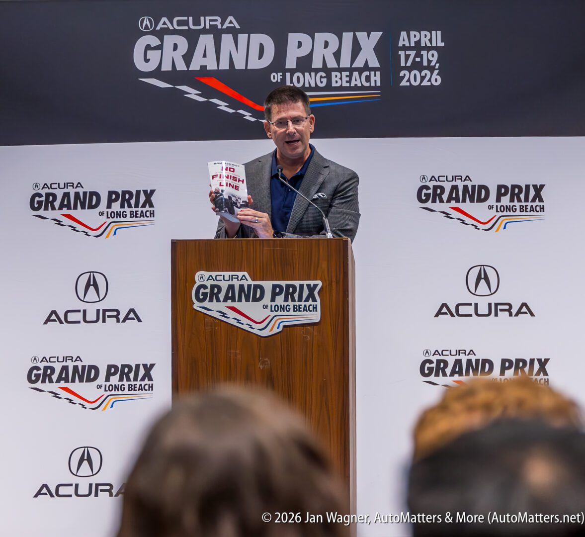 A man speaks at a podium during an Acura Grand Prix of Long Beach event, holding a brochure, with event branding and sponsors visible in the background.