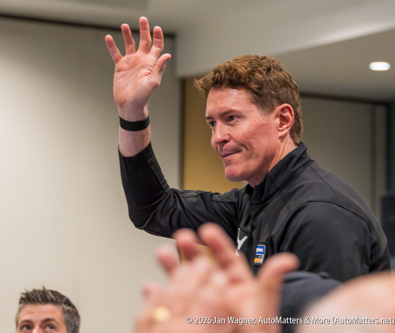 A man in a black jacket stands indoors, raising his right hand in a greeting gesture while people look on.