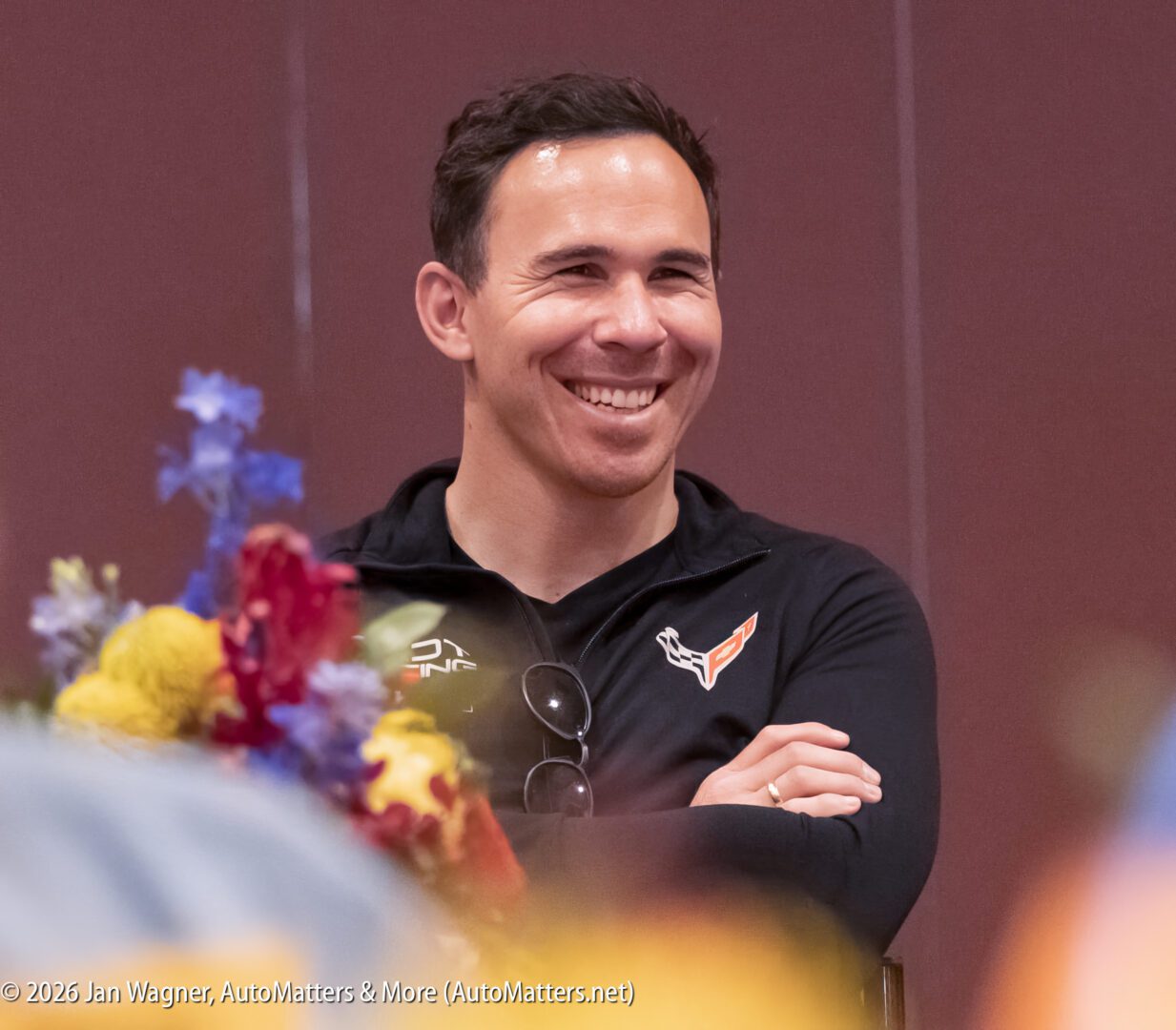 A smiling man wearing a black jacket with a Corvette logo sits indoors, surrounded by blurred flowers in the foreground.