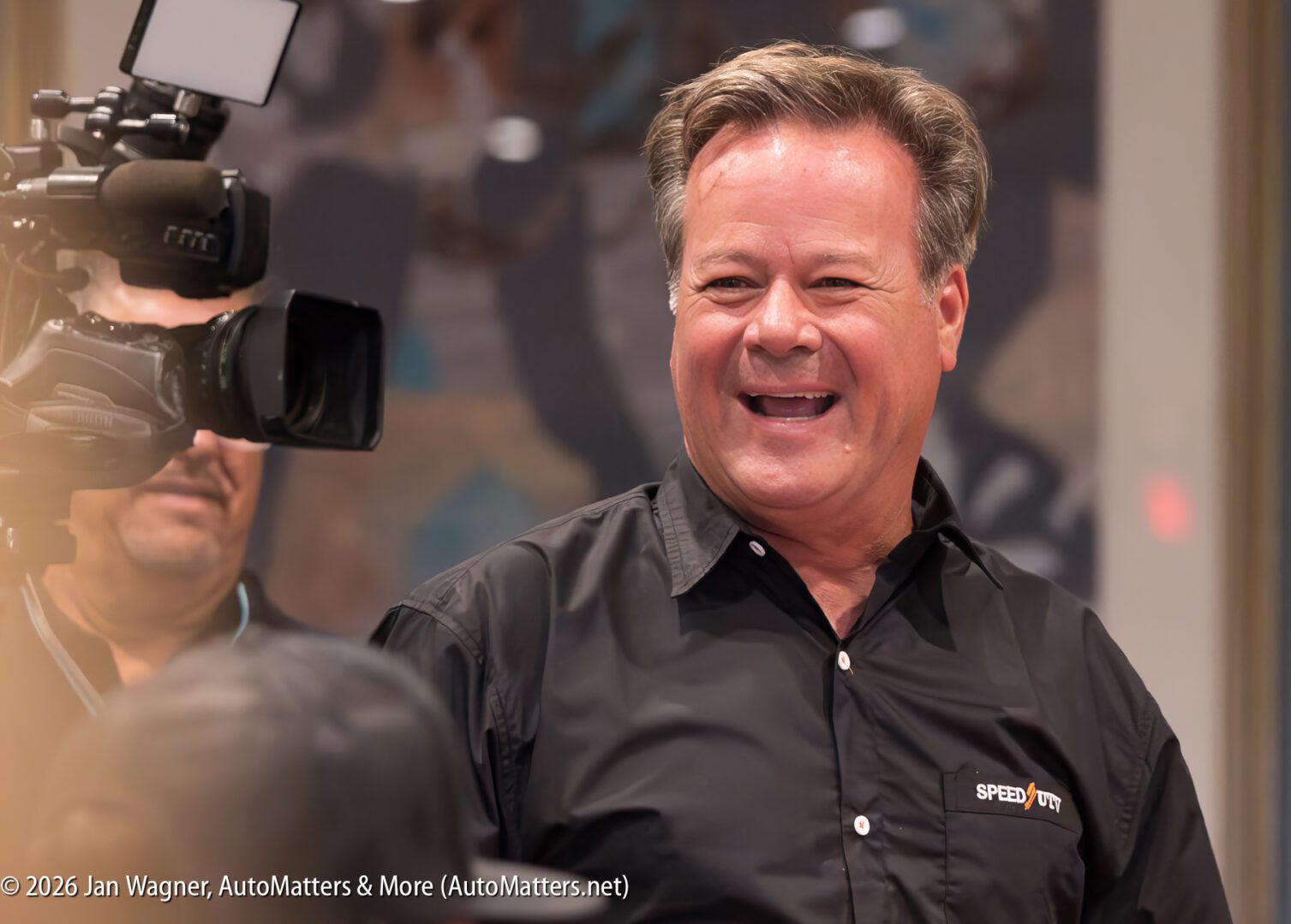 A man in a black SPEED TOUR shirt smiles while being filmed and photographed at an indoor event.