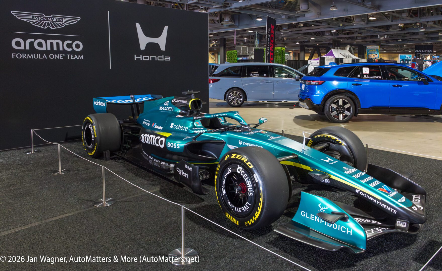 Aston Martin Formula 1 race car on display indoors, surrounded by ropes, with a Honda logo wall and a blue SUV in the background.