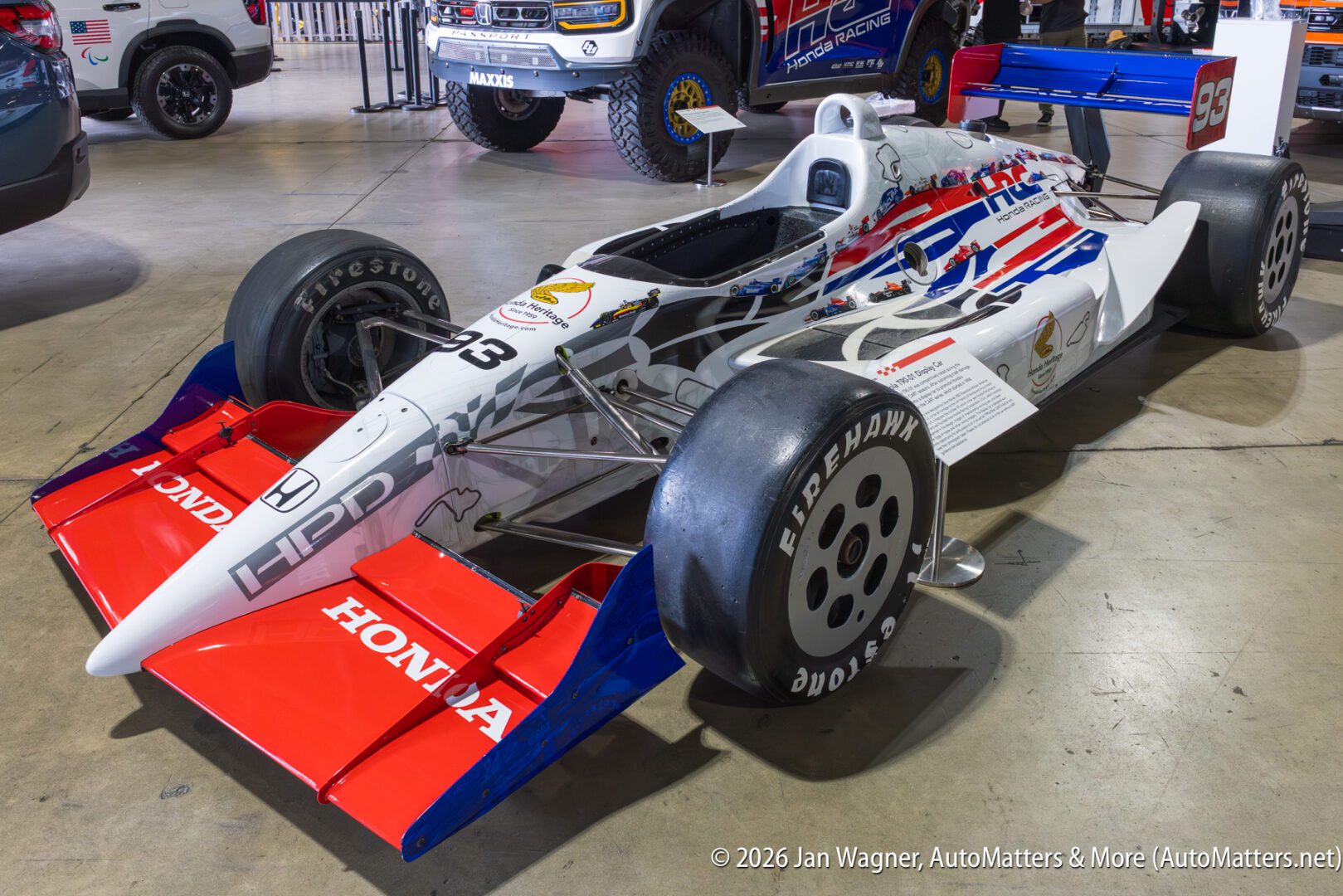 An open-wheel IndyCar race car with red, white, and blue livery is displayed indoors on a polished floor, surrounded by other vehicles.