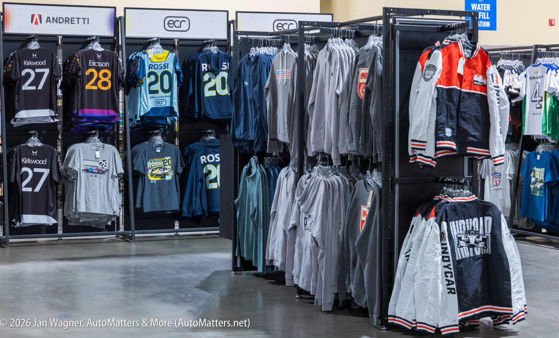 Racks of racing-themed jackets, shirts, and jerseys with drivers' names and numbers on display in a retail setting.