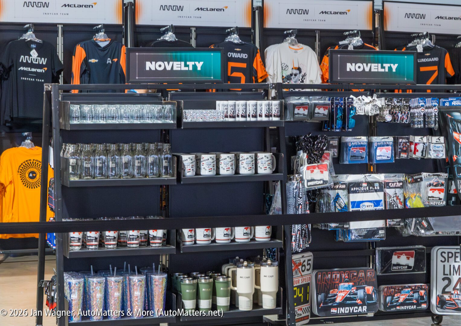 Display shelves filled with McLaren-themed merchandise, including t-shirts, mugs, tumblers, keychains, lanyards, and novelty items, in a retail store setting.