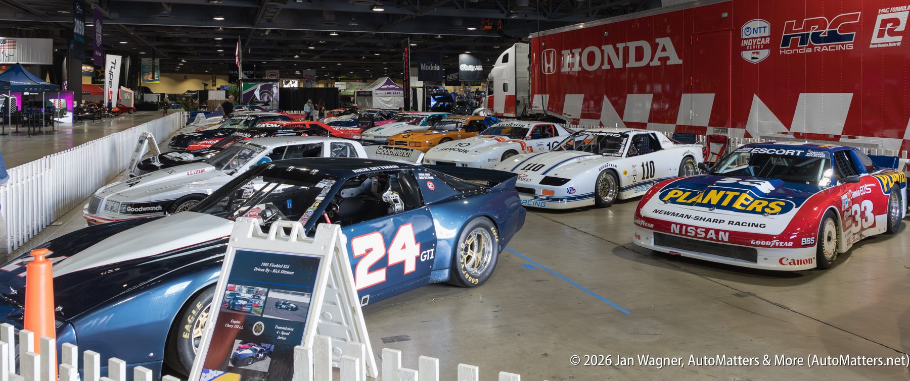 A row of vintage race cars displayed indoors behind a white fence, with a large red Honda trailer and informational signs visible in the background.