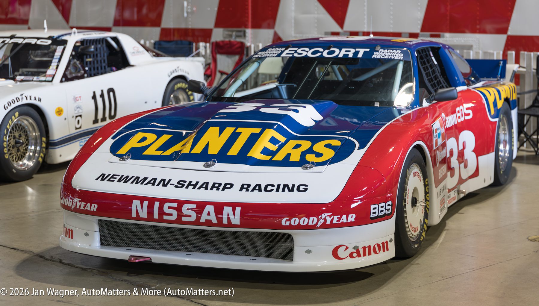 A red, white, and blue Nissan race car with Planters branding and the number 33 on the side, displayed indoors.