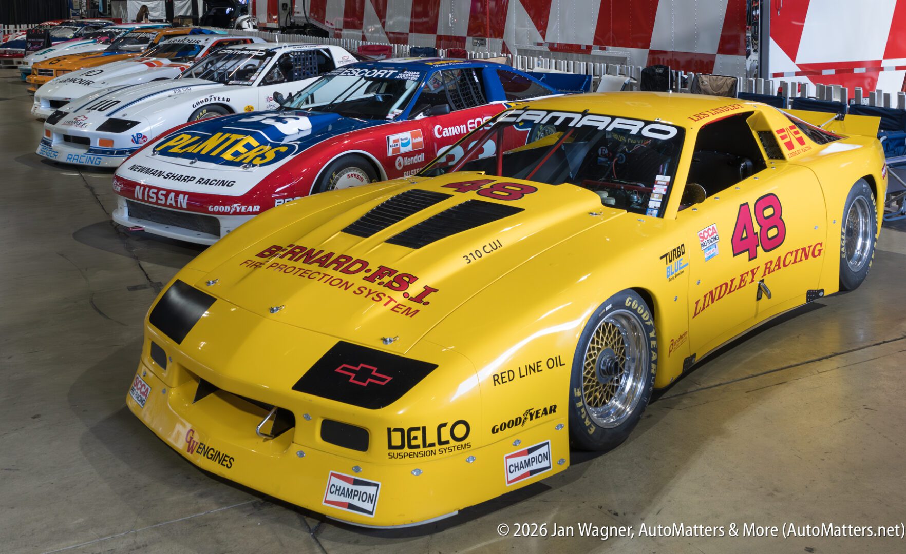 A lineup of vintage race cars indoors, with a prominent yellow #48 Camaro in front, followed by several other cars in various colors and sponsor logos.