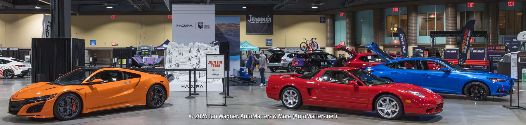 A row of sports cars on display indoors at an auto show, including an orange and a red Acura NSX, with informational booths and banners in the background.