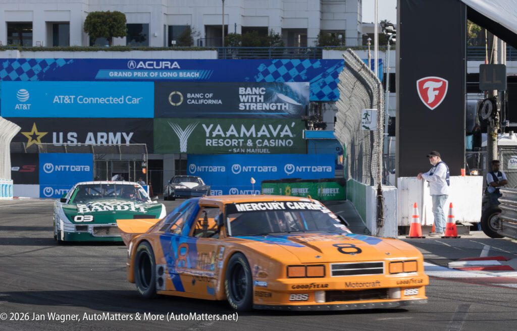 Two vintage race cars, one orange and one green, drive on a city racetrack during a motorsport event, with barriers and sponsor banners visible in the background.