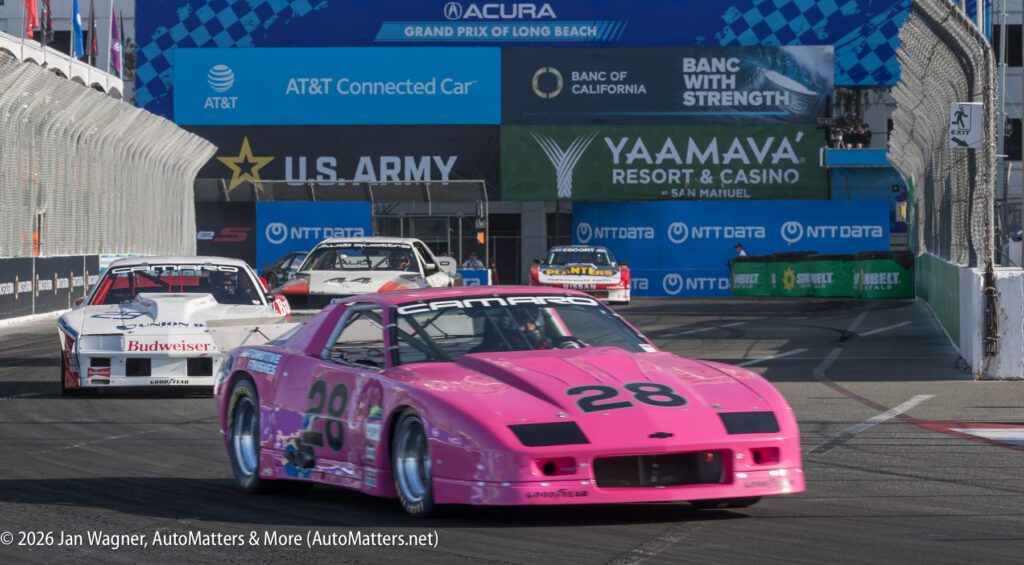 A pink Chevrolet Camaro race car with the number 28 leads a group of race cars on a city street circuit during an auto race event.
