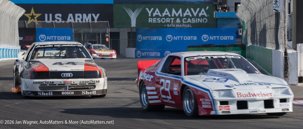 Two vintage race cars, an Audi and a Camaro, compete on a city circuit, passing barriers with visible sponsor logos and advertisements in the background.