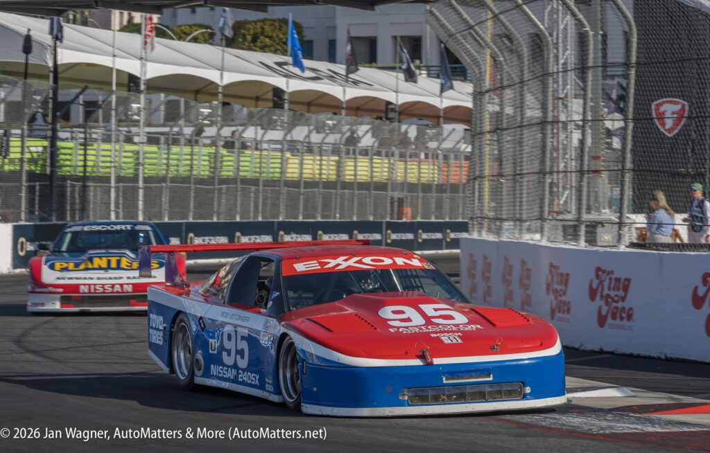 Two Nissan race cars numbered 95 and 33 drive on a closed street circuit during a motorsport event, with grandstands and barriers visible in the background.