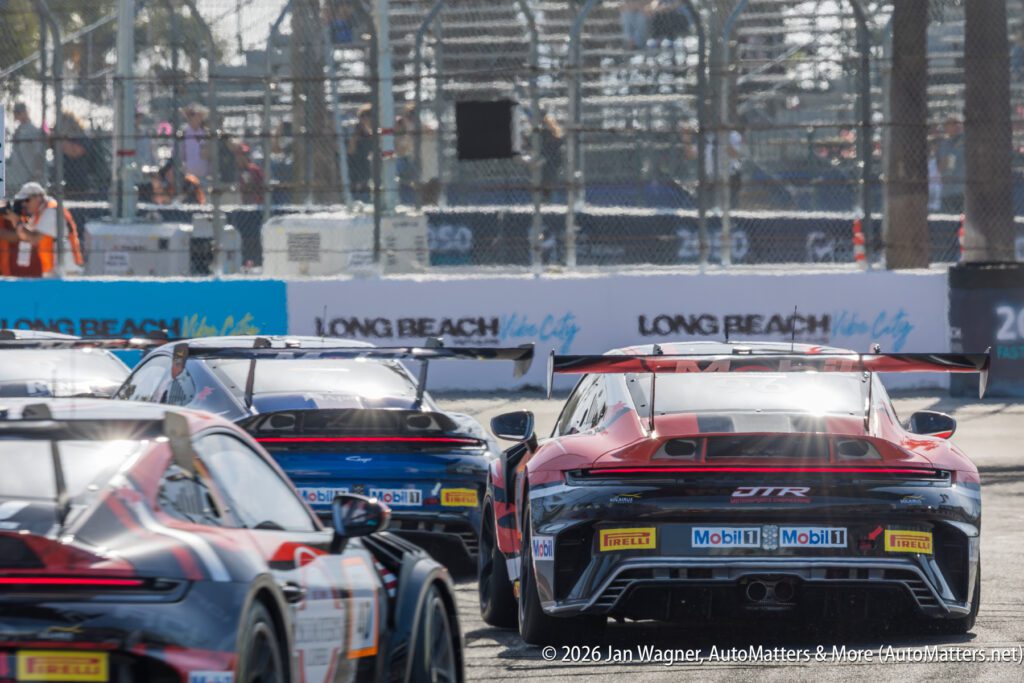 Several racing cars speed on a track at the Long Beach Grand Prix, with barriers and grandstands visible in the background.