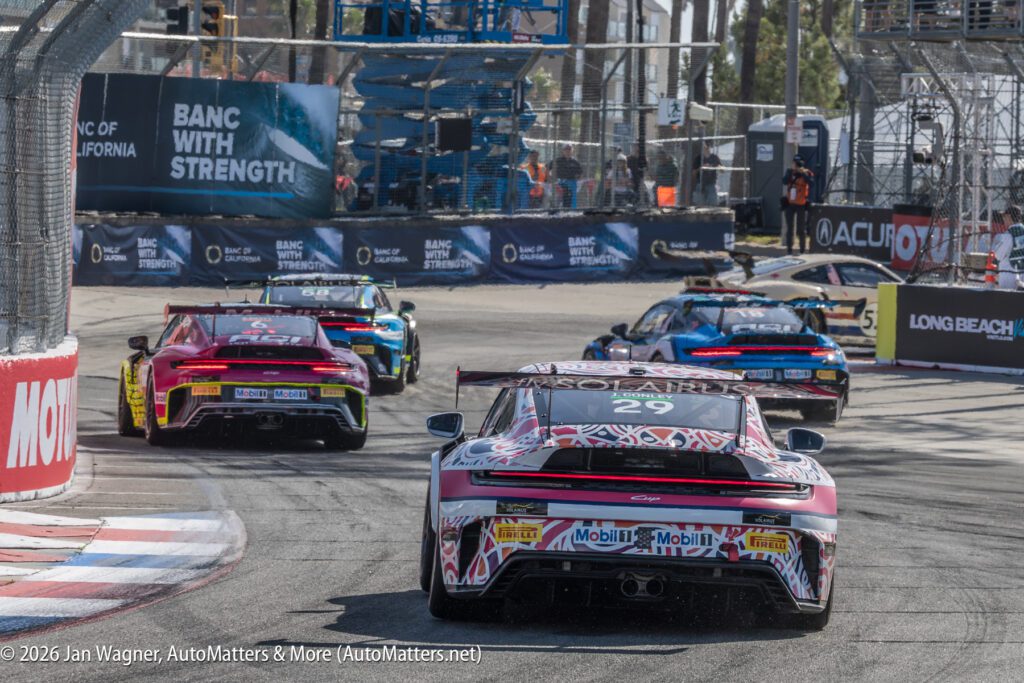 Several race cars navigate a turn on a city street circuit during a motorsports event, with barriers, sponsors, and safety personnel visible in the background.