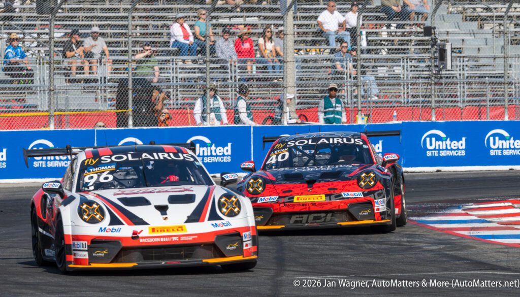 Two Porsche race cars with Solaris branding compete on a street circuit, with spectators watching from grandstands in the background.