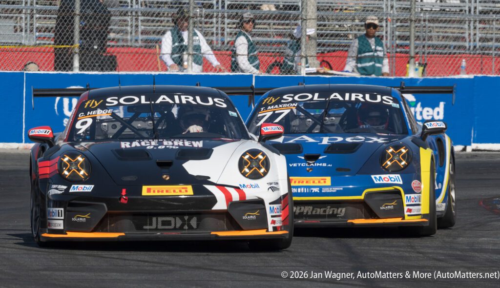 Two Porsche race cars with sponsor logos compete closely on a racetrack, with grandstands and fencing visible in the background.