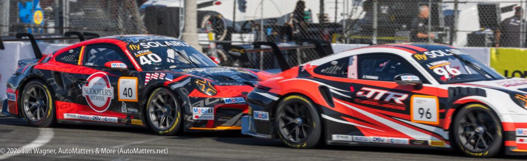 Two sports cars with racing decals and numbers 40 and 96 compete on a race track, passing barriers and spectators in the background.