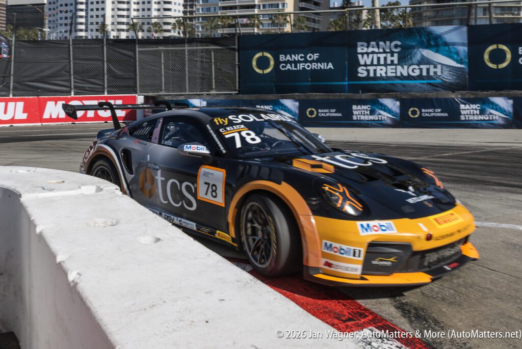 A black and orange race car numbered 78 turns on a street circuit, passing barriers with sponsorship banners and city buildings in the background.