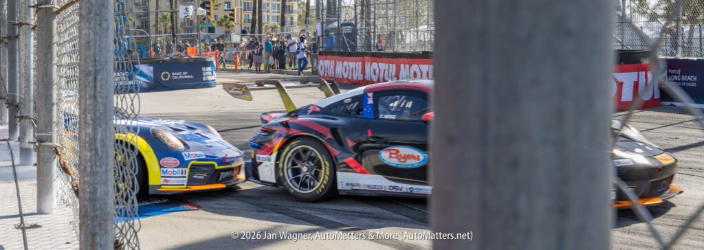 Two race cars turn a corner on a city street circuit, viewed through a chain-link fence with spectators in the background.